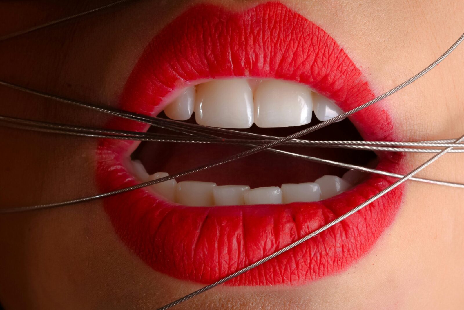 Artistic close-up of red-painted lips with metallic wire crossing the mouth.